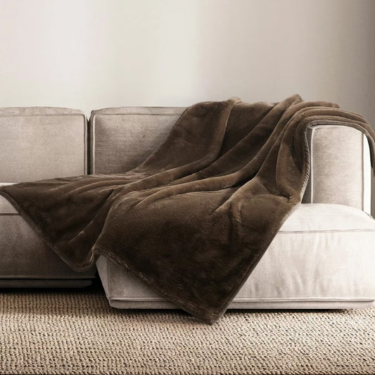 A plush grey Mon Chateau faux fur throw draped casually over the arm of a dark leather sofa in a cozy living room setup.
