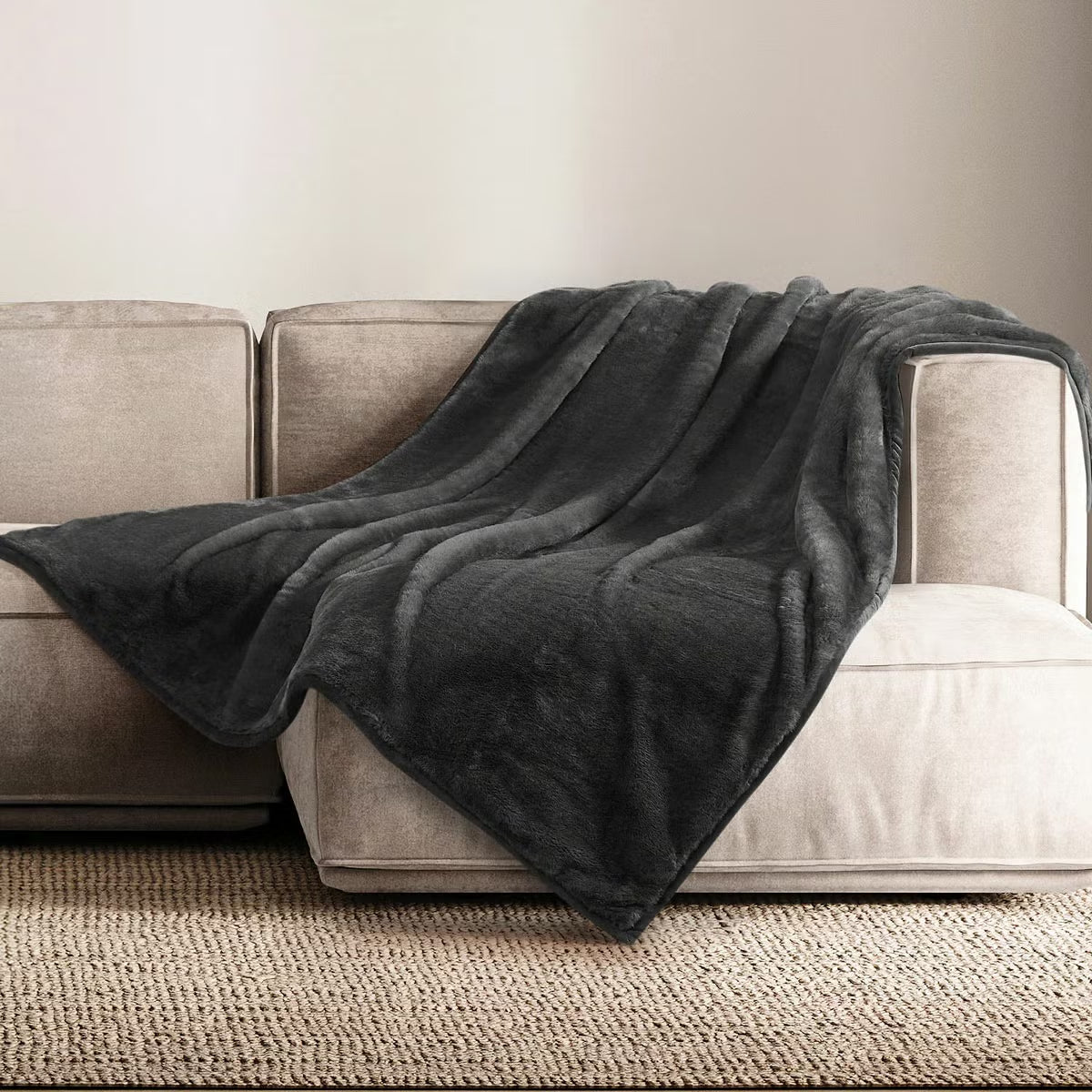 A plush grey Mon Chateau faux fur throw draped casually over the arm of a dark leather sofa in a cozy living room setup.