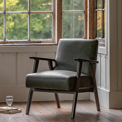 A high-angle close-up of the Gallery Neyland green leather armchair showing its textured top-grain leather seat and solid wood legs in a brightly lit, modern living room.