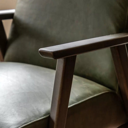 A high-angle close-up of the Gallery Neyland green leather armchair showing its textured top-grain leather seat and solid wood legs in a brightly lit, modern living room.