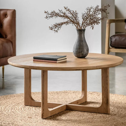 Natural light oak round coffee table featuring a geometric crossover timber base styled on a textured rug in a bright, minimalist modern living room.