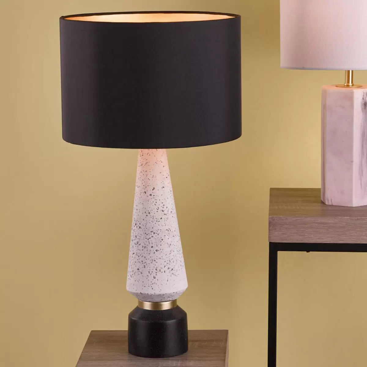 Front view of the Onora table lamp featuring a cylindrical black and gold flecked terrazzo base, topped with a black faux silk drum shade on a concrete sideboard.
