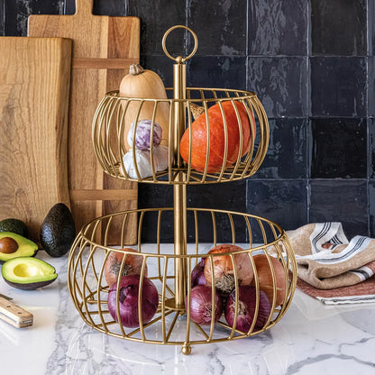 Over & Back 2-Tier brushed gold metal basket displayed on a kitchen counter filled with fresh fruit.