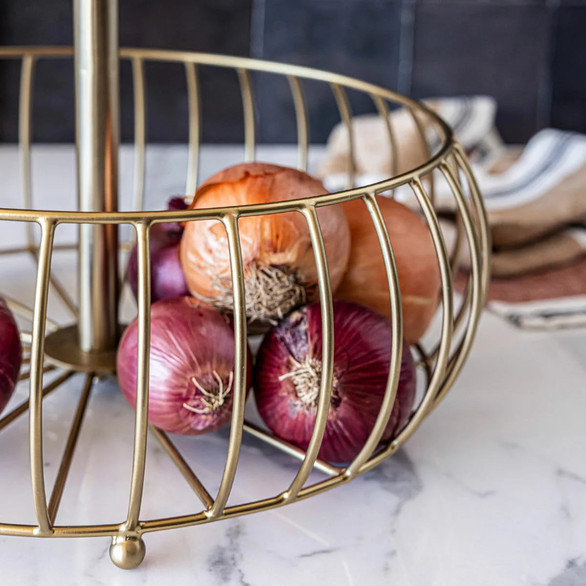 Over & Back 2-Tier brushed gold metal basket displayed on a kitchen counter filled with fresh fruit.