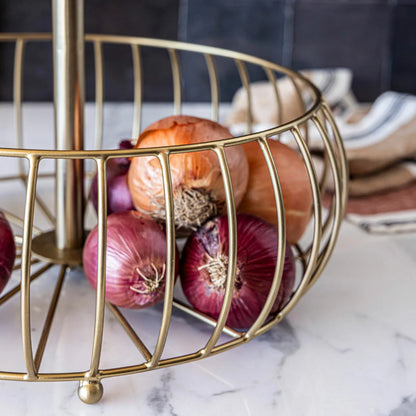 Over & Back 2-Tier brushed gold metal basket displayed on a kitchen counter filled with fresh fruit.