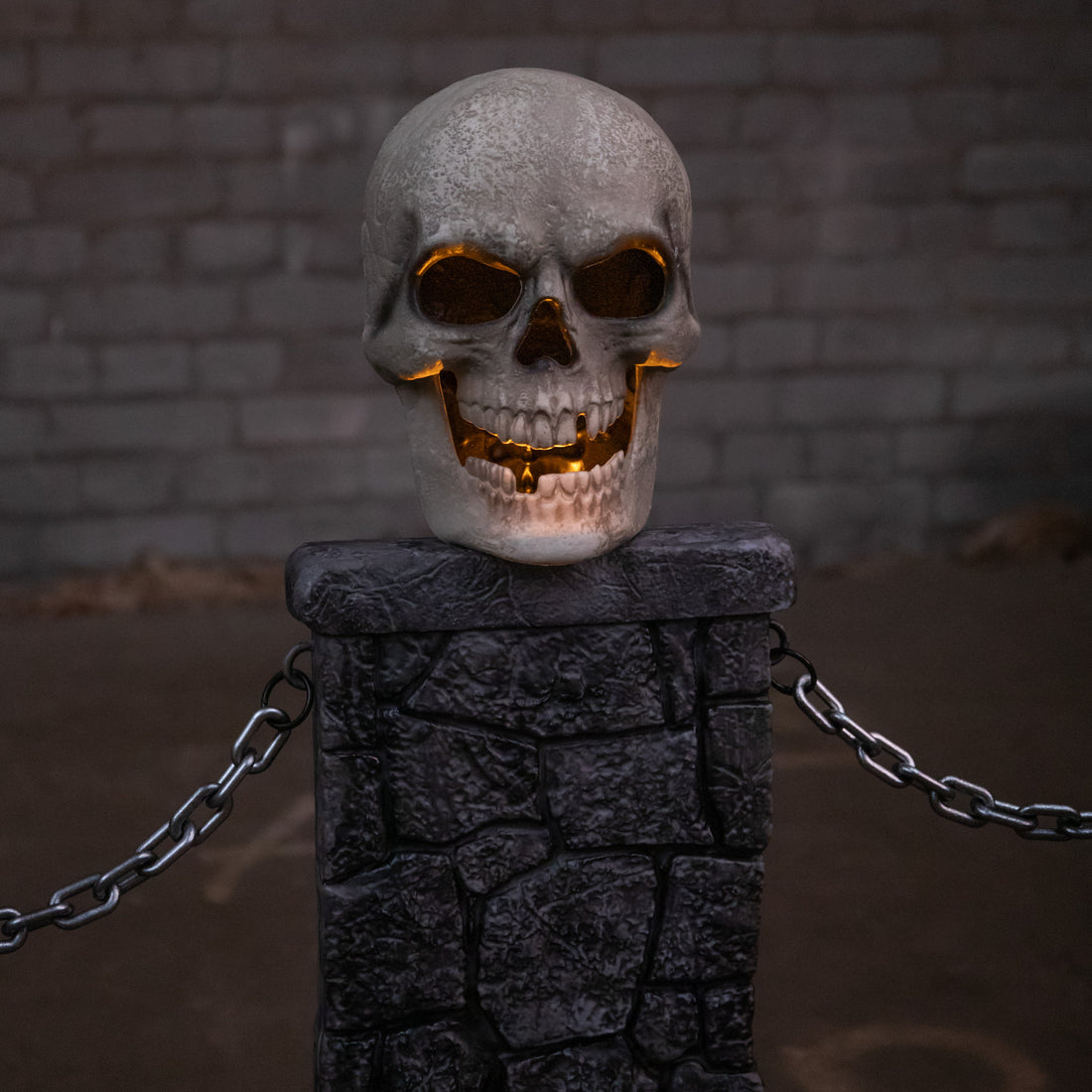 Pair of grey stone-effect Halloween skull fence sets with glowing LED eyes and linking chains anchored into a dark front lawn at dusk.