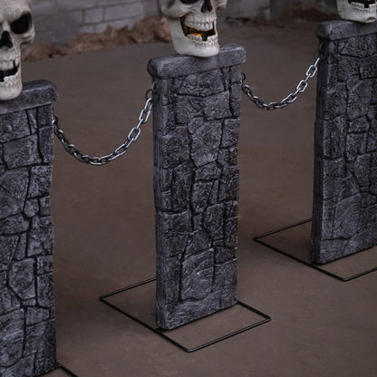 Pair of grey stone-effect Halloween skull fence sets with glowing LED eyes and linking chains anchored into a dark front lawn at dusk.