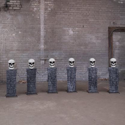 Pair of grey stone-effect Halloween skull fence sets with glowing LED eyes and linking chains anchored into a dark front lawn at dusk.