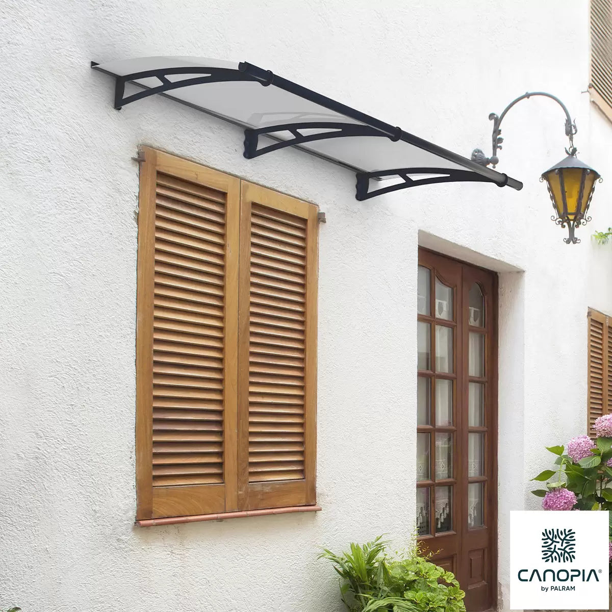 Angle view of a clear Palram Canopia Aquila 1500 door canopy with dark grey steel arms mounted over a residential front entrance.