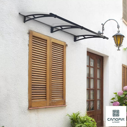 Angle view of a clear Palram Canopia Aquila 1500 door canopy with dark grey steel arms mounted over a residential front entrance.