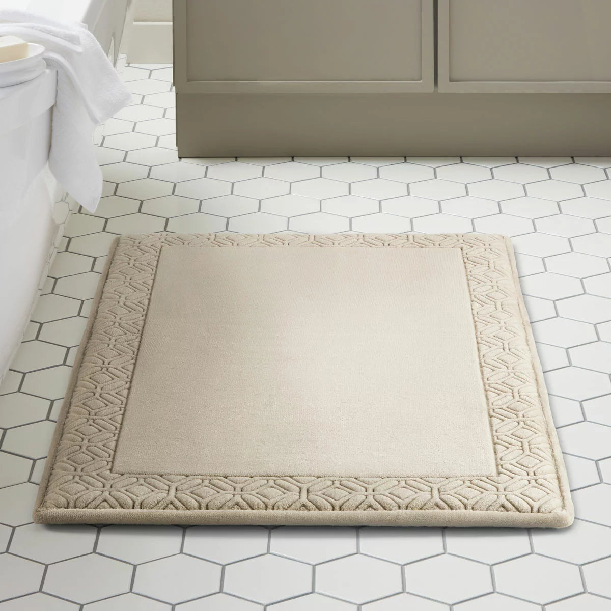 Top view of the Paramount taupe memory foam plush bath mat showing leaf-bordered stitched fabric and soft plush texture on a neutral tile floor.