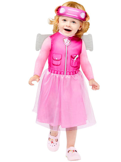 Front view of a toddler wearing the pink PAW Patrol Skye dress with attached aviator wings and character headband standing in a colorful nursery setting.