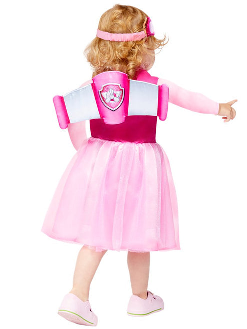 Front view of a toddler wearing the pink PAW Patrol Skye dress with attached aviator wings and character headband standing in a colorful nursery setting.