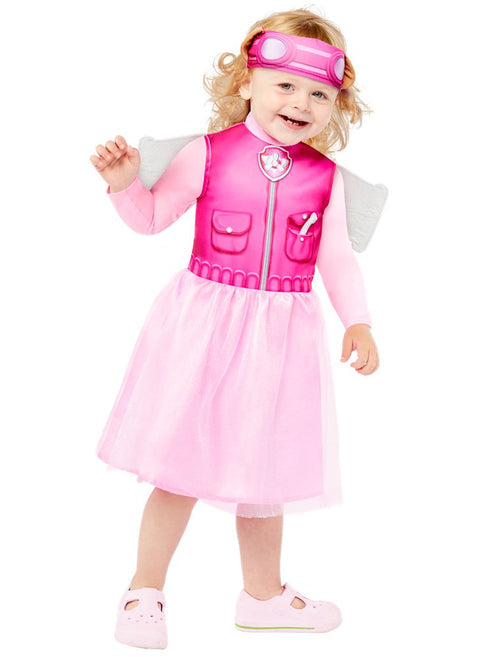 Front view of a toddler wearing the pink PAW Patrol Skye dress with attached aviator wings and character headband standing in a colorful nursery setting.