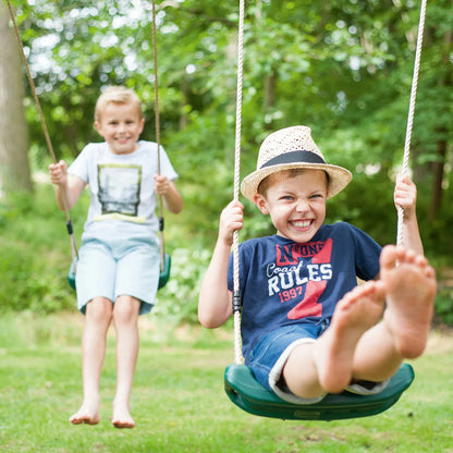 A Plum Ultimate premium wooden swing set standing in a lush green garden, featuring an assembled frame with a baby seat and trapeze attached, plus safety mats underneath.