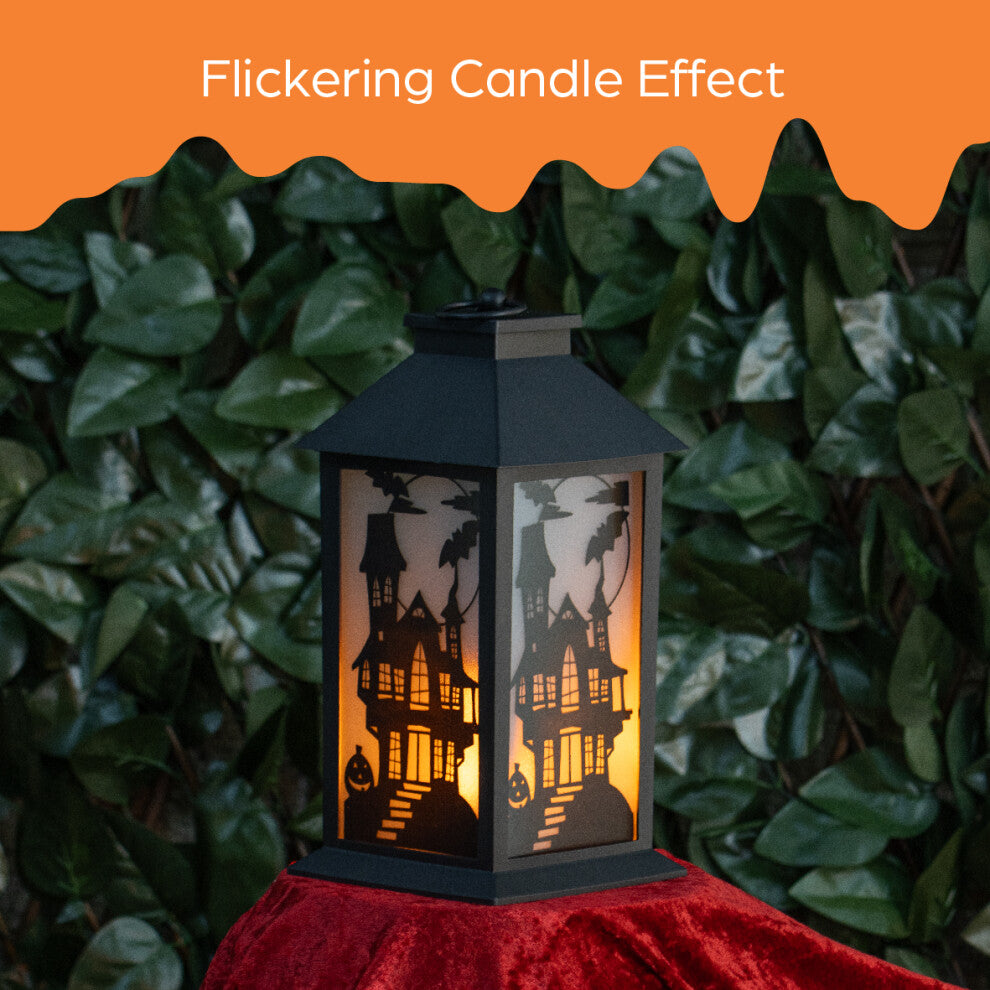 A 30.5cm black Victorian-style Global Gizmos LED Halloween lantern featuring a flickering orange candle effect and haunted house panels sitting on a wooden surface.