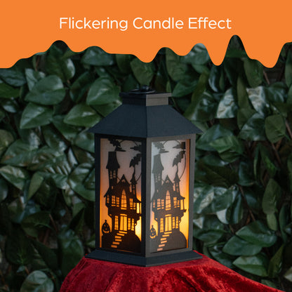 A 30.5cm black Victorian-style Global Gizmos LED Halloween lantern featuring a flickering orange candle effect and haunted house panels sitting on a wooden surface.