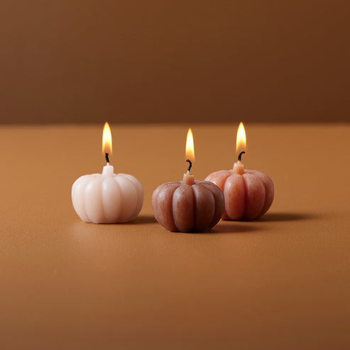 Three orange pumpkin shaped candles with detailed ribbed wax textures displayed on a dark wooden table beside rustic autumn leaves.