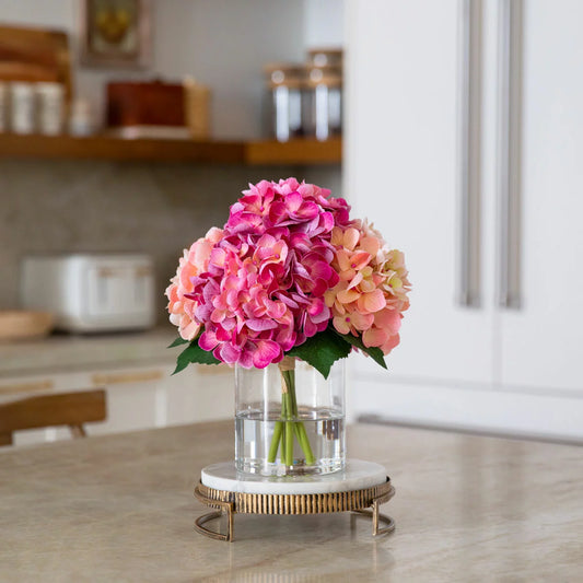 A real-touch artificial hydrangea arrangement with lush, vibrant petals inside a clear glass cylinder vase featuring a realistic faux water finish.