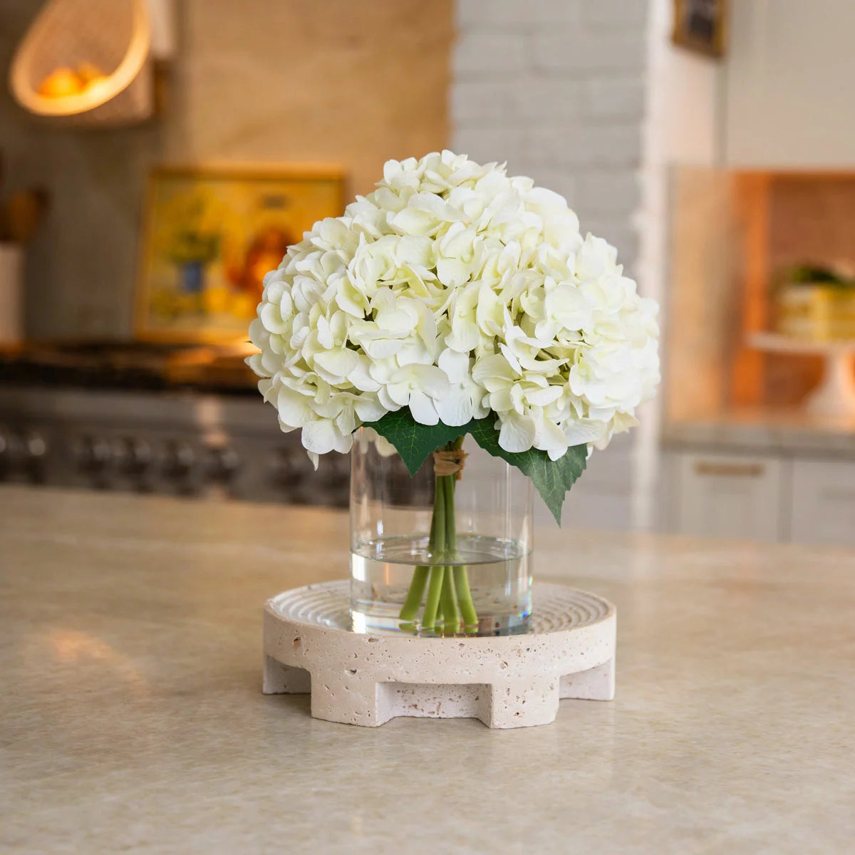 A real-touch artificial hydrangea arrangement with lush, vibrant petals inside a clear glass cylinder vase featuring a realistic faux water finish.