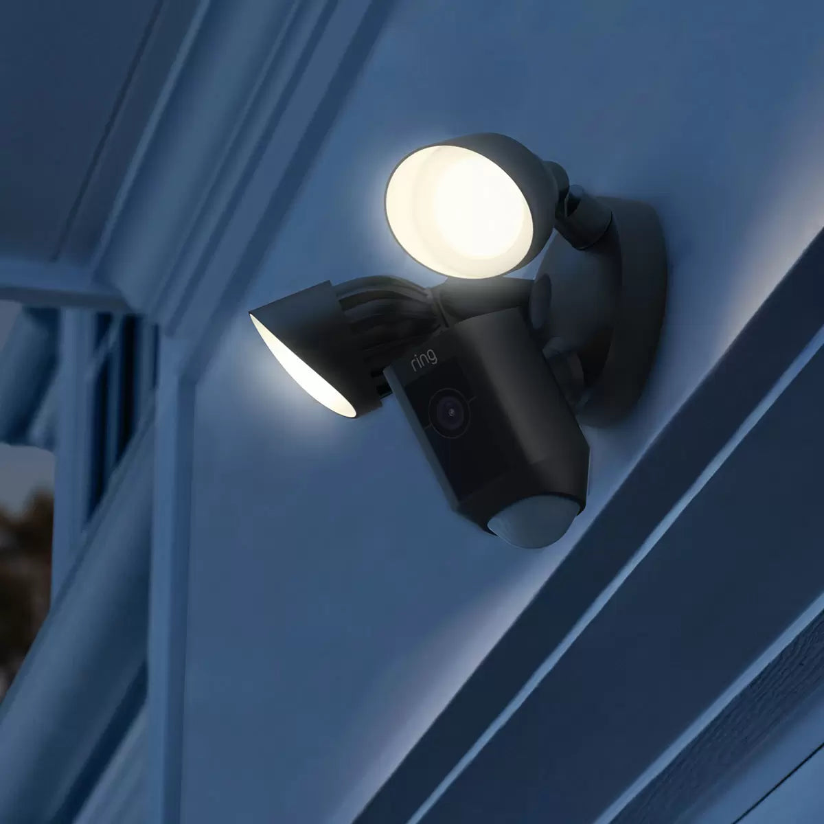 A wall-mounted matte black Ring Floodlight Cam Plus showing twin adjustable LED panels, a central camera housing, and a matching Chime Pro plugged into an indoor wall socket.