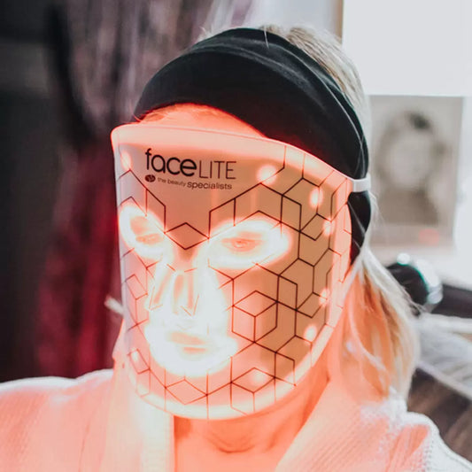 A flexible, white Rio faceLite silicone LED face mask shown glowing with soft red interior lights, resting flat on a contemporary vanity table next to its USB charging cord.
