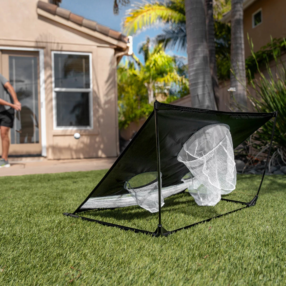 Black SKLZ Quickster Chipping Net set up on a green lawn with three distinct target holes and a golf ball resting near the bottom collection slot.