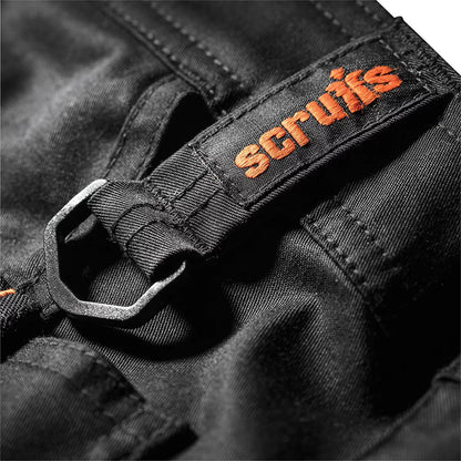 Side angle shot of a tradesman wearing black Scruffs Trade Flex Trousers with loaded holster pockets and integrated CORDURA knee pads on a dusty indoor construction site.