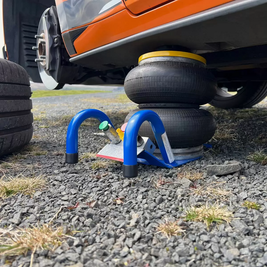 Side profile of the blue Selson HBNXLO 2-bellow hand-held air jack resting on a concrete workshop floor, highlighting its robust rubber bellows, wide support base, and long metal control handle.
