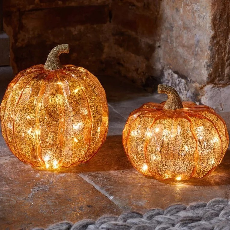 A set of two light-up LED pumpkin ornaments in different sizes glowing warmly on a dark rustic wooden autumn table.