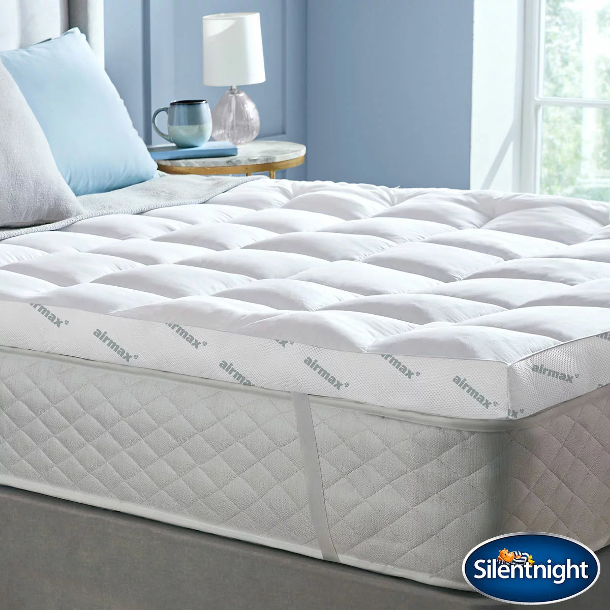 Silentnight Airmax 1000 Mattress Topper fitted on a modern bed with visible breathable mesh trim and elastic corner straps in a bright bedroom setting.