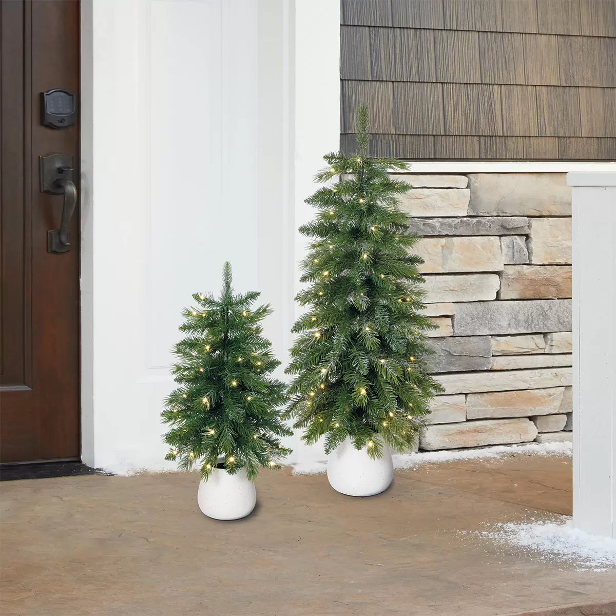 Pair of 2ft and 3ft silver glitter potted artificial trees with glowing warm white twinkling LED lights sitting on a white porch threshold.