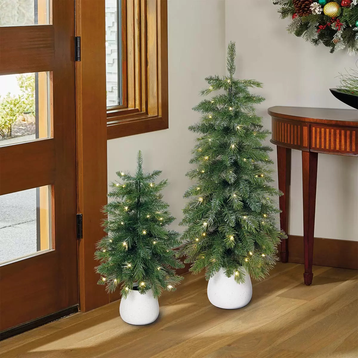 Pair of 2ft and 3ft silver glitter potted artificial trees with glowing warm white twinkling LED lights sitting on a white porch threshold.