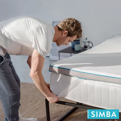 Close-up angled view of a white Simba Hybrid mattress topper featuring a breathable mesh cover and blue border detailing laid over a wooden bed frame in a light-filled bedroom.