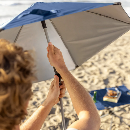 Low-profile blue folding Sport-Brella beach chair with breathable mesh back, padded armrest cup holder, and attached UPF 50+ shade umbrella set up on sunny ocean sand.