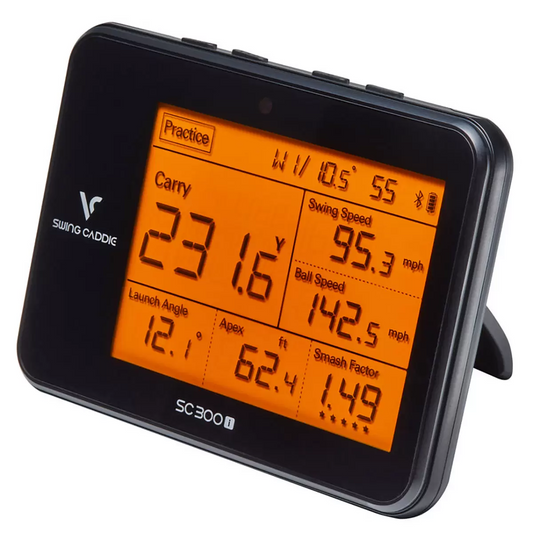Sleek charcoal grey Swing Caddie SC300i launch monitor standing upright on a synthetic driving range mat, displaying green numeric yardages on its lit screen with a driver resting beside it.