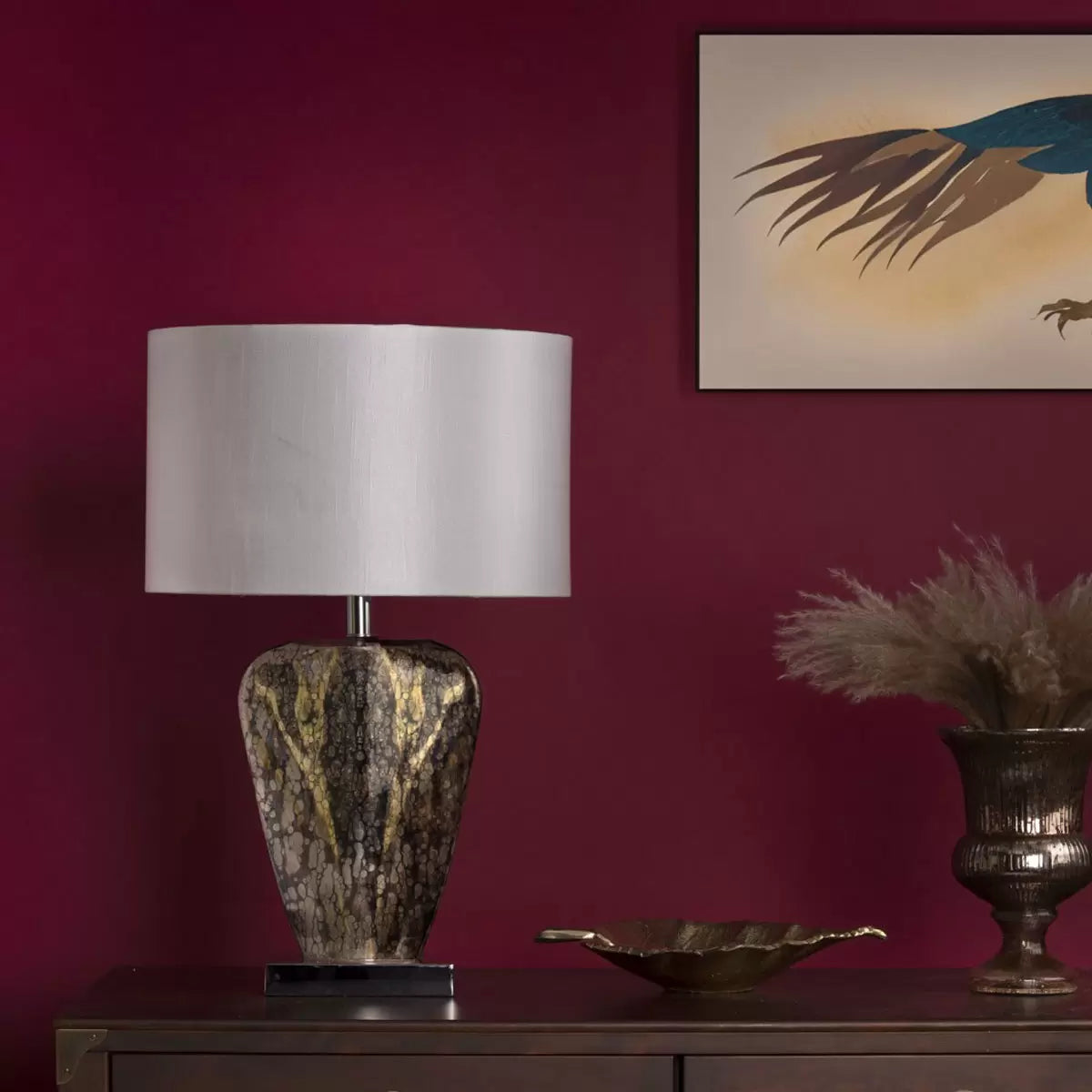 A close-up profile shot of the Syracuse table lamp showcasing its reflective, antique silver-and-gold mercury glass base and textured cream faux silk shade set against a neutral, wood-paneled wall.