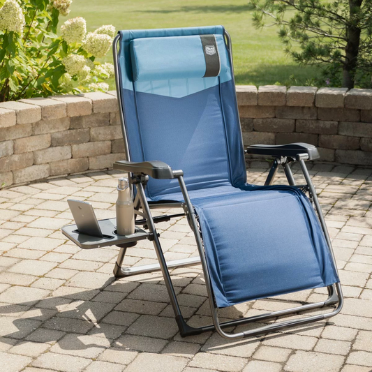 A dark padded Timber Ridge Zero Gravity Folding Lounger fully extended on a sunny outdoor patio, showcasing its open side table with a cup holder slot and the attached adjustable headrest pillow.