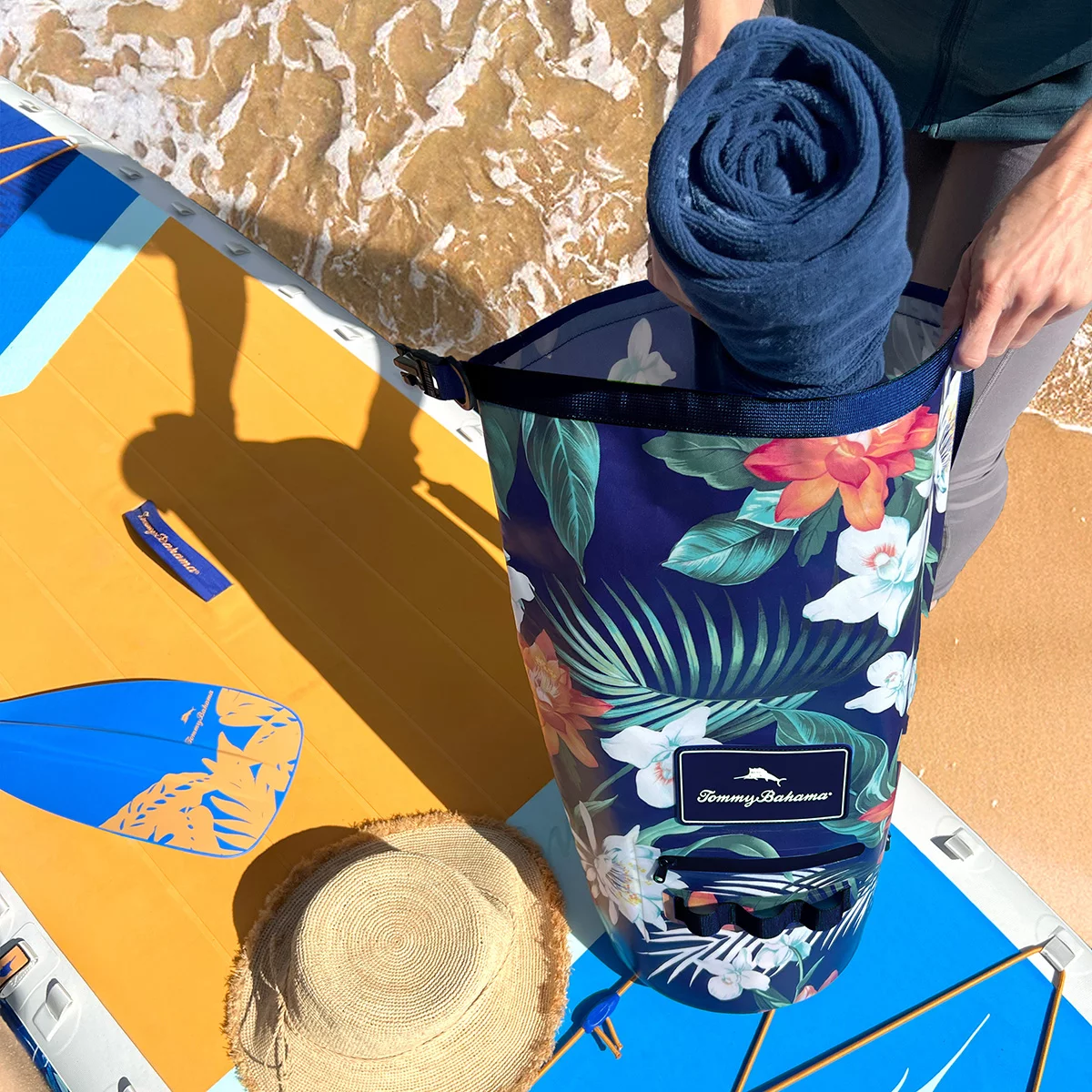 Vibrant tropical leaf print Tommy Bahama 20L TPU-coated waterproof dry bag with rolled buckle top and shoulder strap sitting on a wet wooden boat dock.