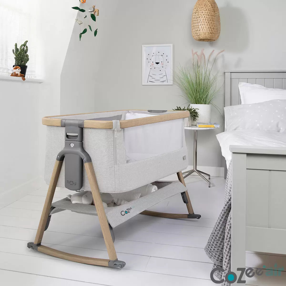 Side view of the Tutti Bambini CoZee Air bedside crib in oak and silver finish showing dual mesh windows, mattress, and detachable rocking arches inside a neutral nursery.