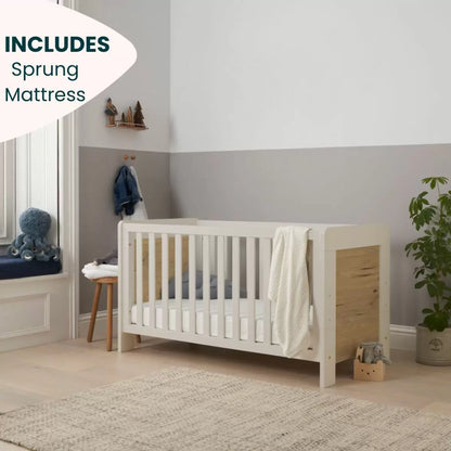 Front angle of the Tutti Bambini Alba Cot Bed in White Sand and Distressed Oak finish showing the included sprung mattress inside a brightly lit nursery setting.