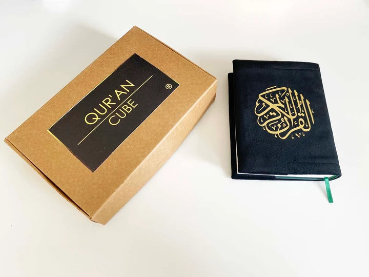 Angle view of a black soft velvet covered 13-line Quran with gold Arabic logo next to its matching velvet holder inside an open presentation gift box.