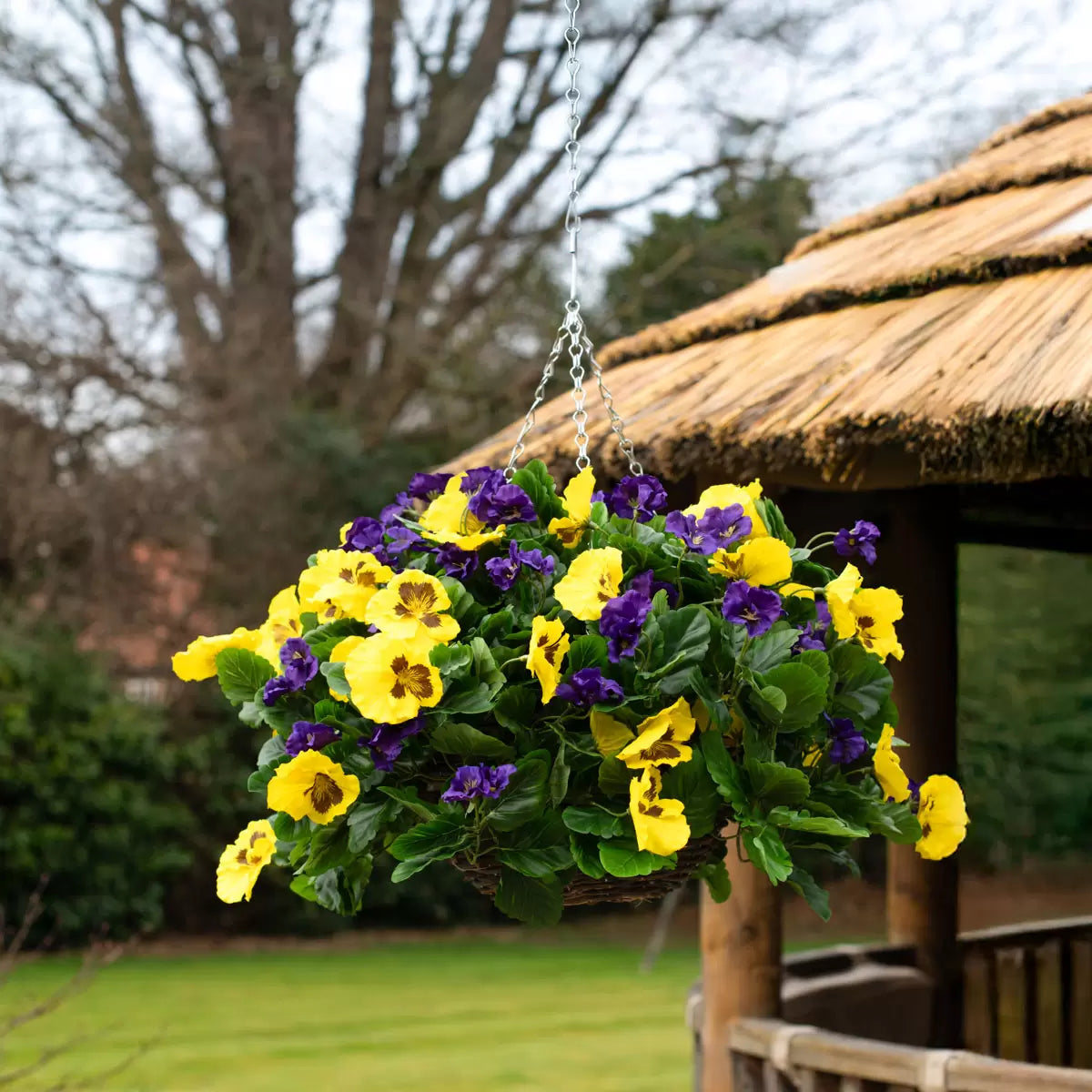 A vibrant artificial flower hanging basket with lush, colourful blooms and green foliage suspended by a sleek metal chain against a clean, neutral wall.