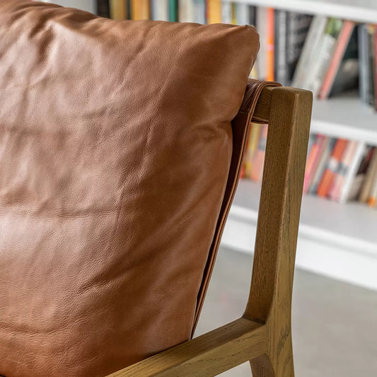 A close-up angled shot of the Gallery Burela Lounge Chair highlighting its vintage brown leather creases, plush seat cushion, and natural grain of the light ash wood legs.