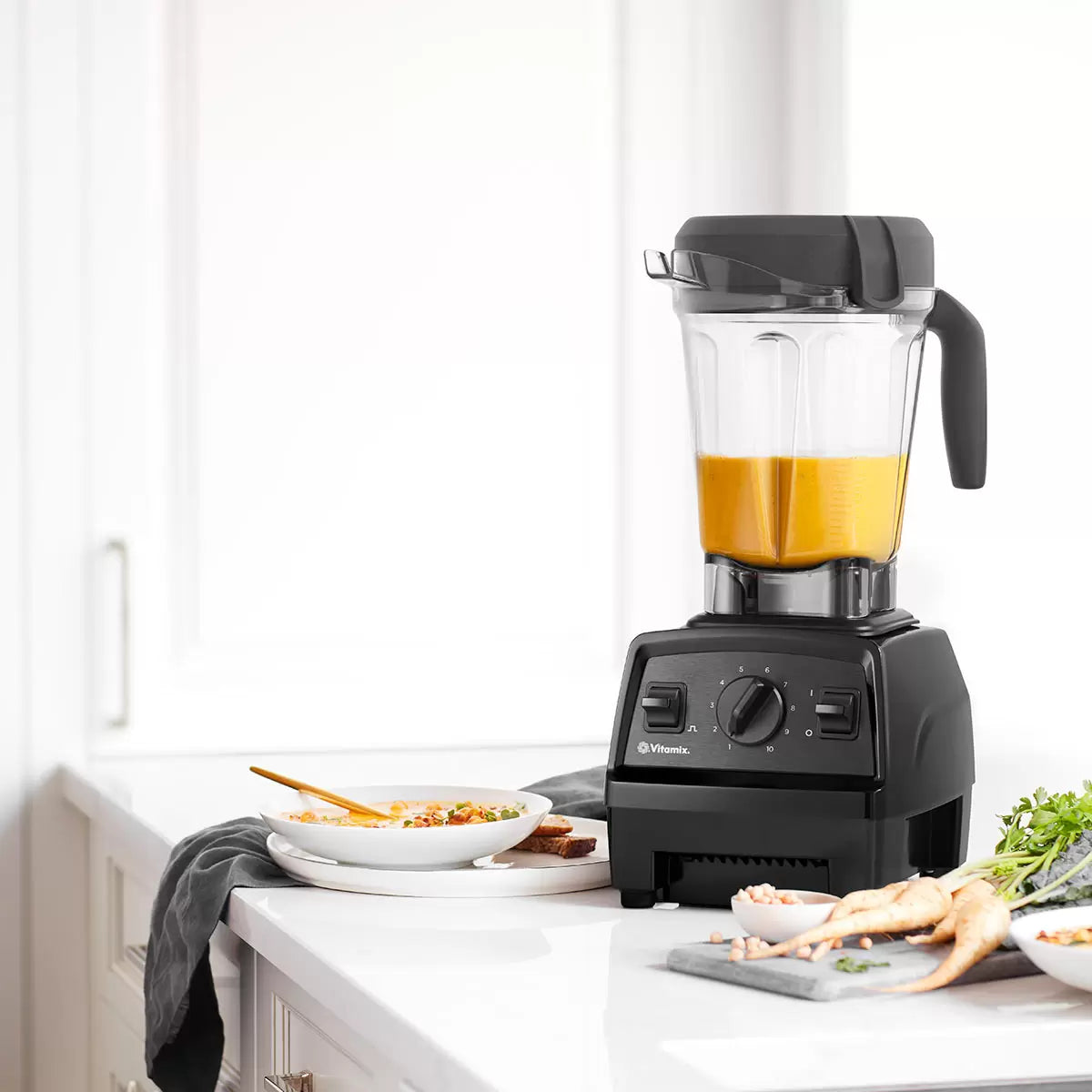 A sleek black Vitamix E320 blender base with its 2L container and tamper sitting on a polished stone kitchen countertop.