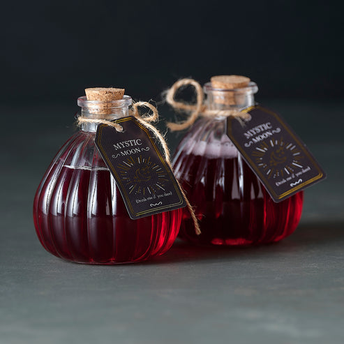 Pair of small clear glass Witch's Brew potion bottles with vintage distressed labels sitting on a dark wooden surface.