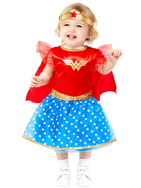 Baby and toddler girl wearing a red, blue, and gold Wonder Woman costume with an attached blue cape and soft fabric headband tiara on a neutral background.