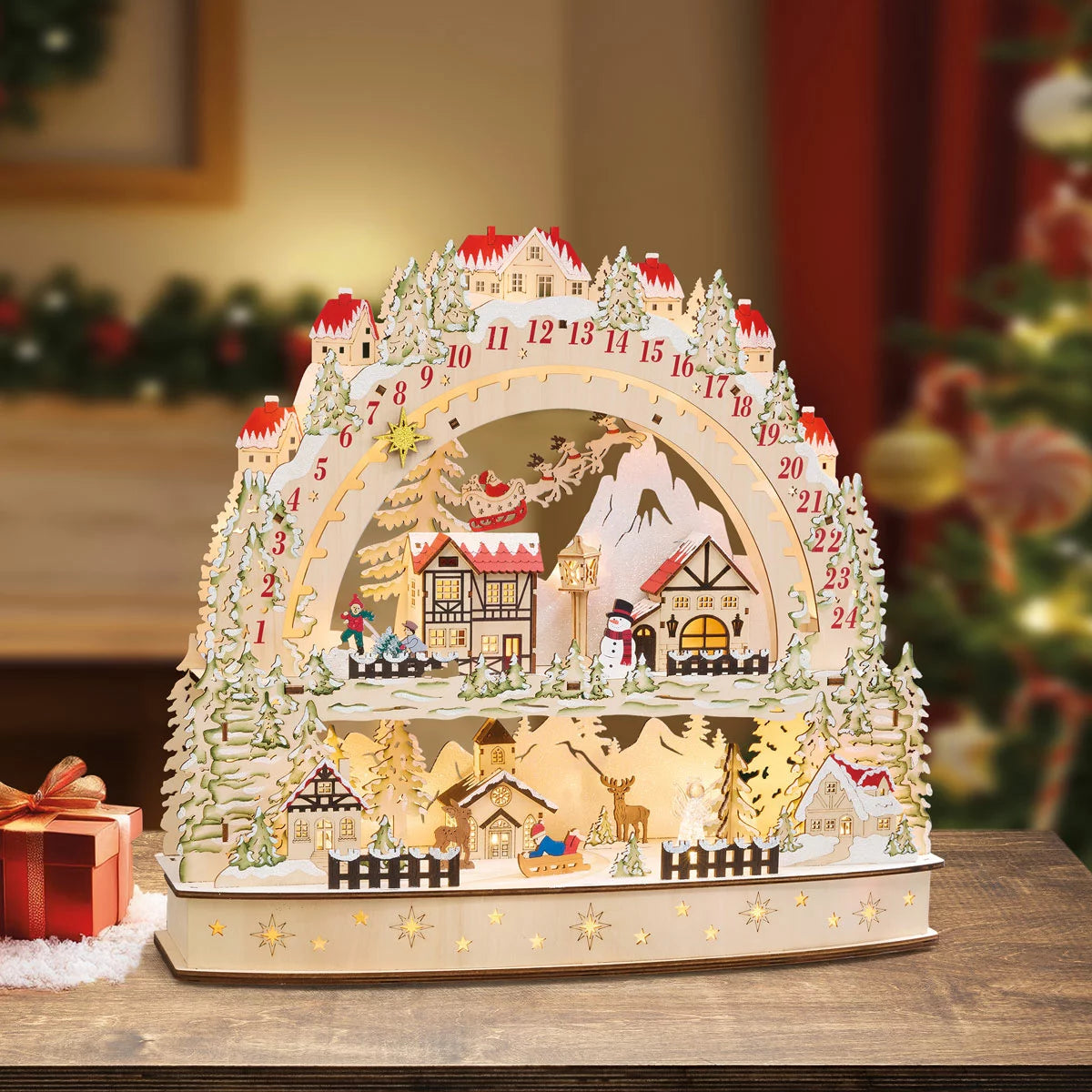 Front view of a laser-cut wooden alpine village advent calendar illuminated by warm internal LED lights on a wooden table.