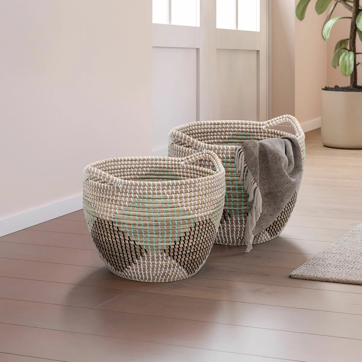 Pair of two nesting handwoven natural brown seagrass storage baskets with integrated handles arranged on a light wood floor in a brightly lit living room.