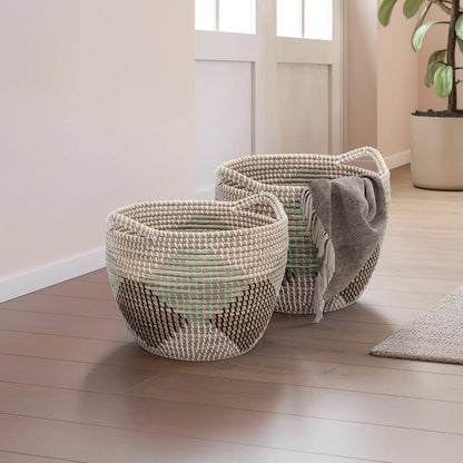 Pair of two nesting handwoven natural brown seagrass storage baskets with integrated handles arranged on a light wood floor in a brightly lit living room.